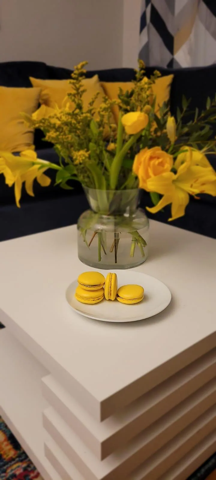 Four bright yellow lemon macarons on a white plate in front of yellow flowers
